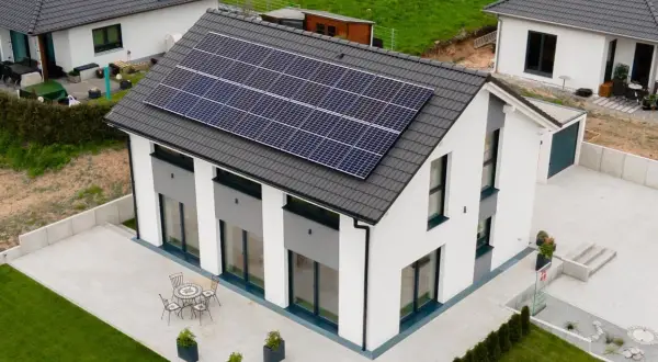 Nowoczesny biały dom z panelami słonecznymi na dachu, otoczony zielonym trawnikiem i patio.